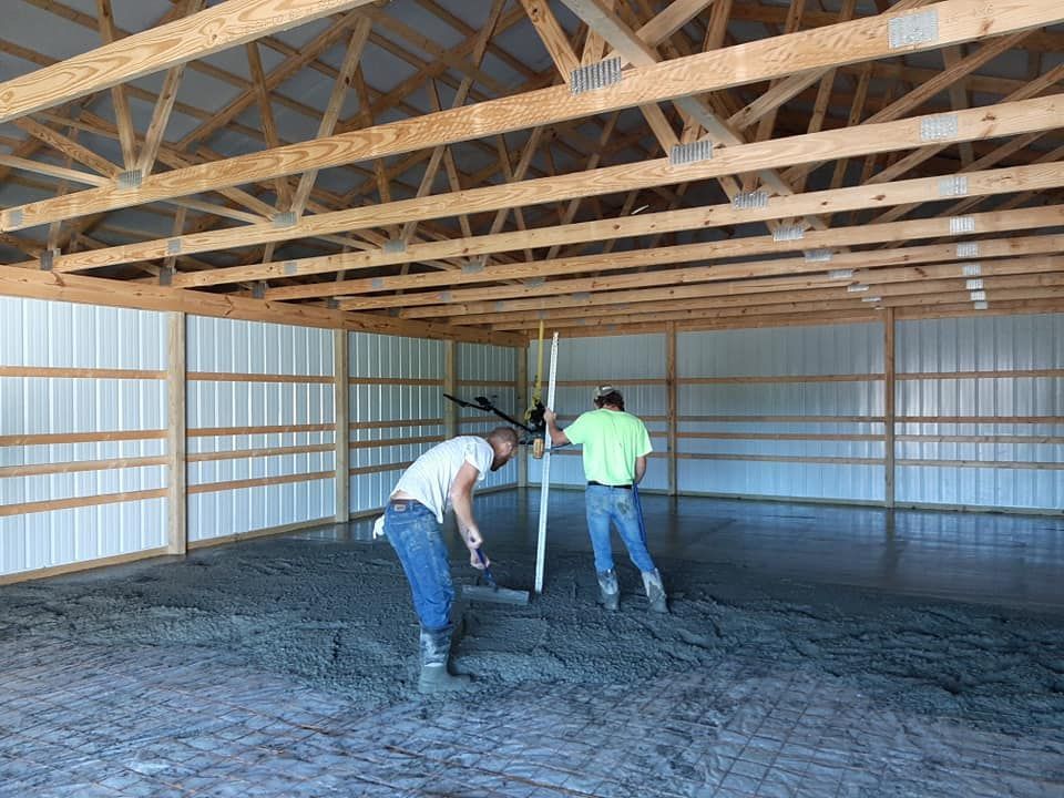 Two workers in a pole barn use tools to level fresh concrete on the floor.