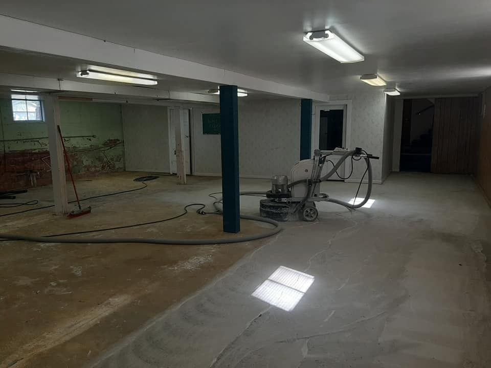 A basement under renovation with a concrete floor, structural pillars, and a floor sander in the middle of the room.