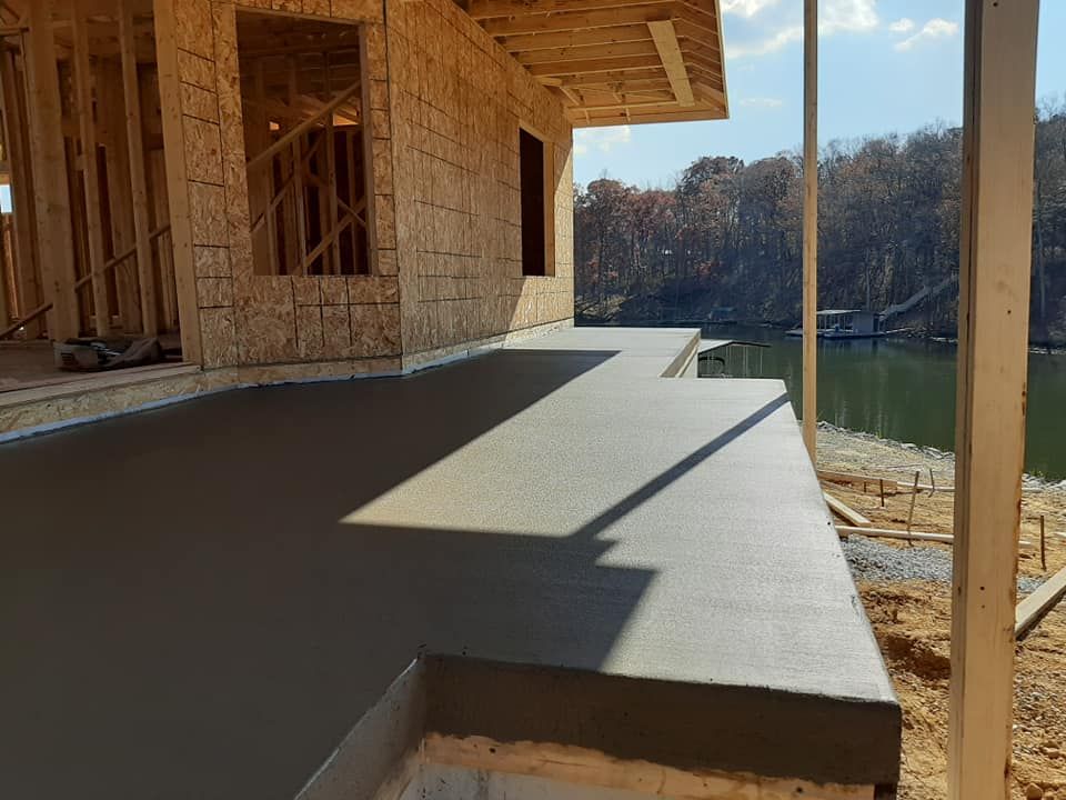 Newly poured concrete patio beside a wooden house frame overlooking a river on a sunny day.