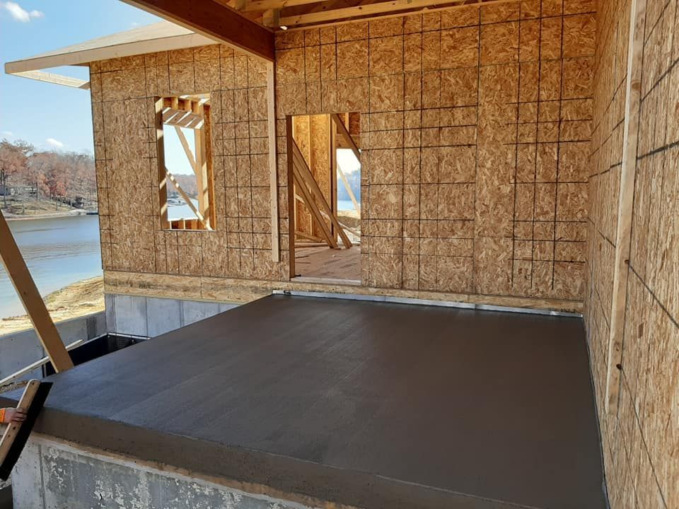 A newly poured concrete porch attached to a wood-framed house under construction overlooking a body of water.