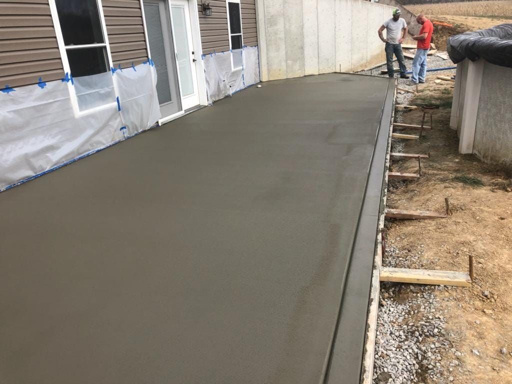 Two people stand near a newly poured concrete patio alongside a house foundation and exposed construction forms.