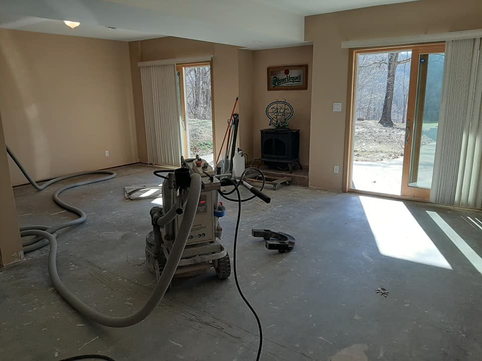 A commercial dust-collection sander sits on a bare subfloor in a sunlit room with sliding glass doors and a wood stove.