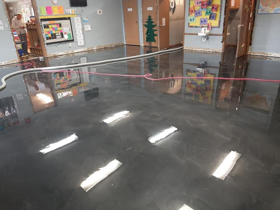 Flooded classroom with dark standing water reflecting the ceiling lights and a person standing in a doorway.