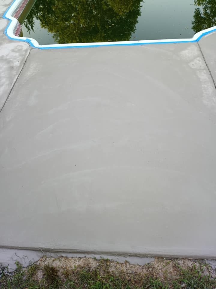 A newly poured concrete slab sits beside a swimming pool, bordered by grass and a blue-tiled edge.