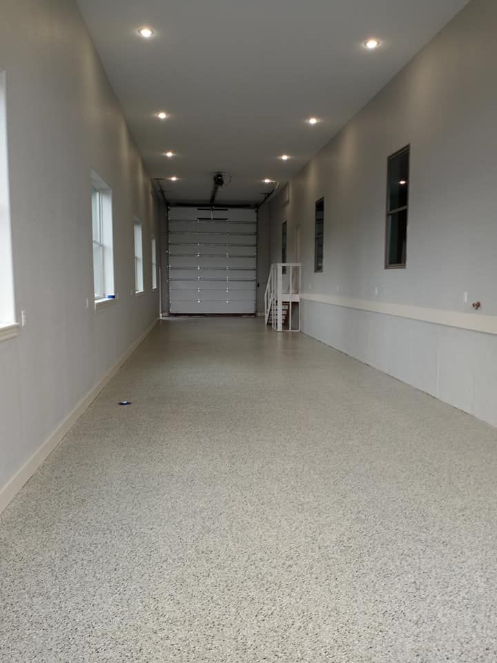 A long, narrow room with white walls, a speckled epoxy floor, recessed ceiling lights, and a closed white garage door.