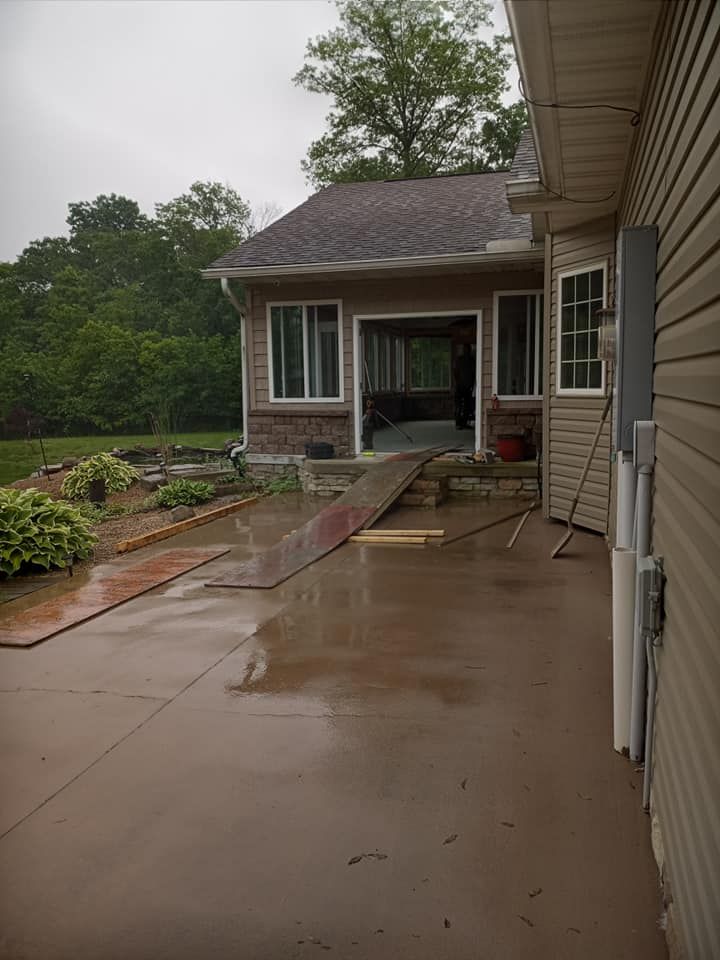 A wet concrete patio leads to a doorway with a makeshift ramp leading into an open room of a tan-sided house.