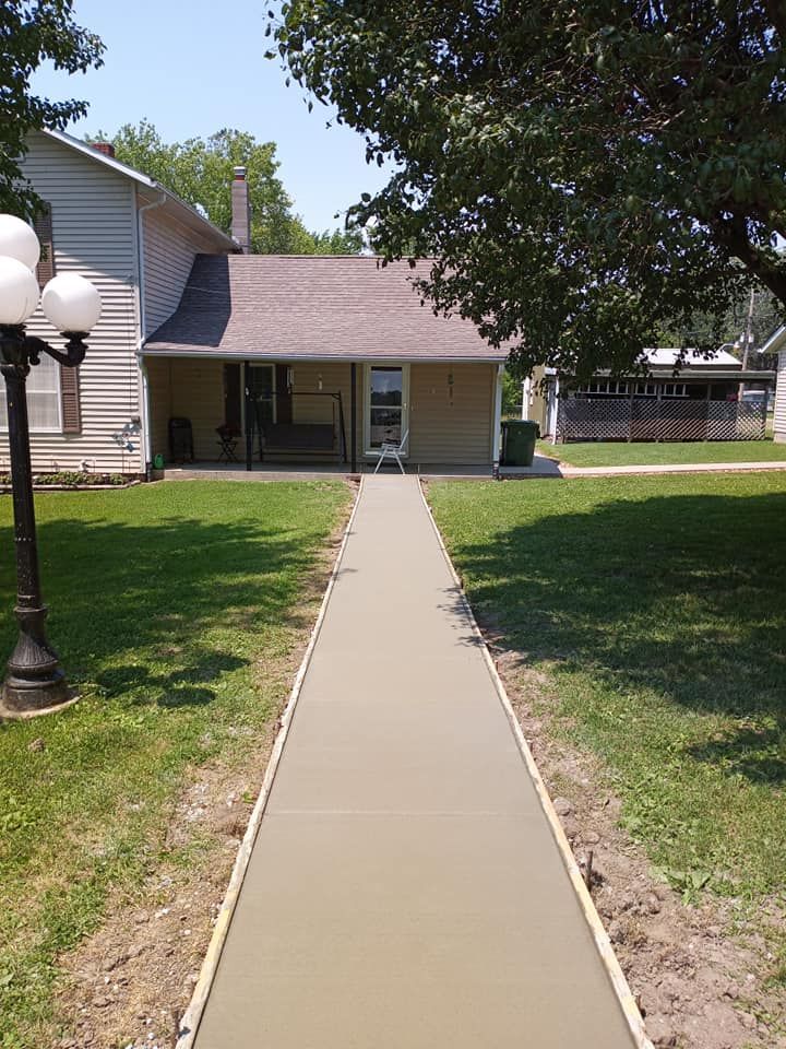 A new concrete sidewalk leads to the covered entrance of a beige house on a sunny day with a lamp post in the foreground.