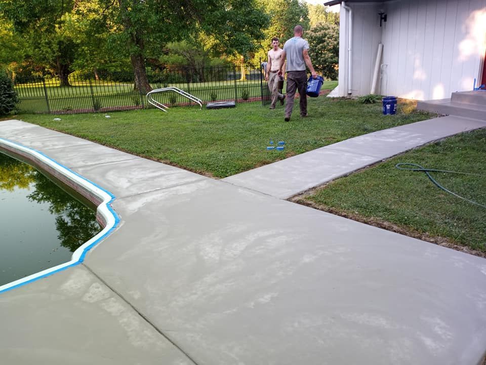 Two people carry buckets across a grassy backyard near a freshly poured concrete patio and a swimming pool.