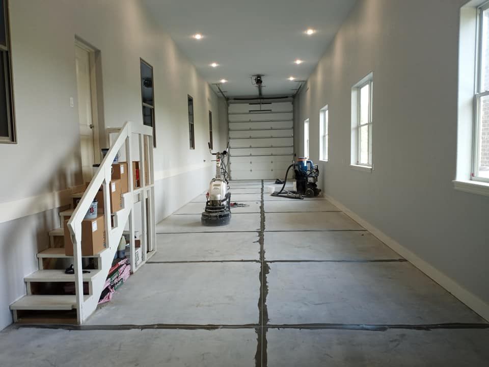 An interior view of a long garage with light gray walls, a concrete floor with visible joint lines, and a wooden staircase.