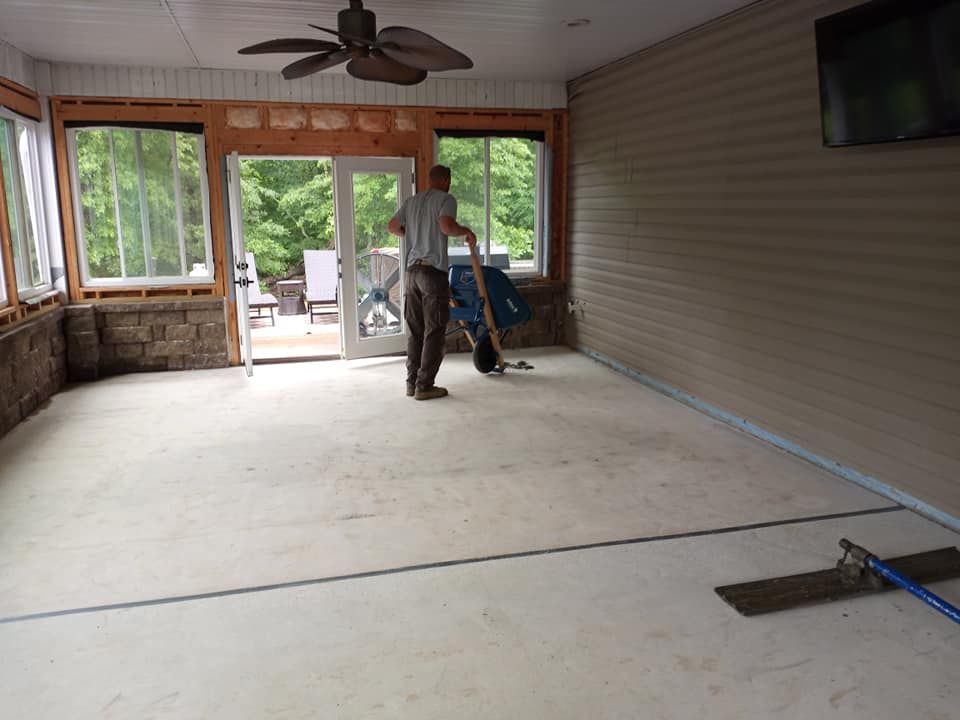 A person using a floor float to smooth wet concrete inside an unfinished sunroom with stone and siding walls.