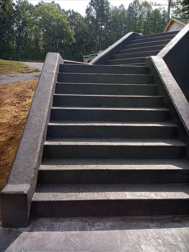 A wide set of dark gray concrete outdoor stairs with side railings, ascending toward a wooded area.