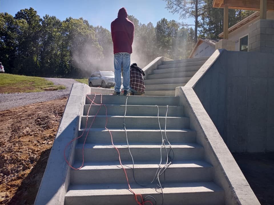 Two people stand on new concrete outdoor stairs at a construction site, with dust in the air and electrical cords draped.
