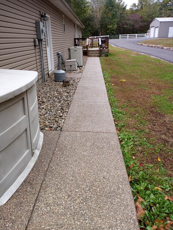 A concrete walkway leads along the side of a tan-sided house toward a wooden deck, with a gravel strip and grassy area.