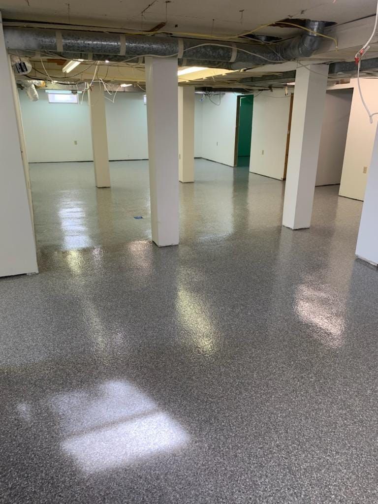 A freshly finished basement with grey speckled epoxy flooring, white support columns, and an exposed ceiling.