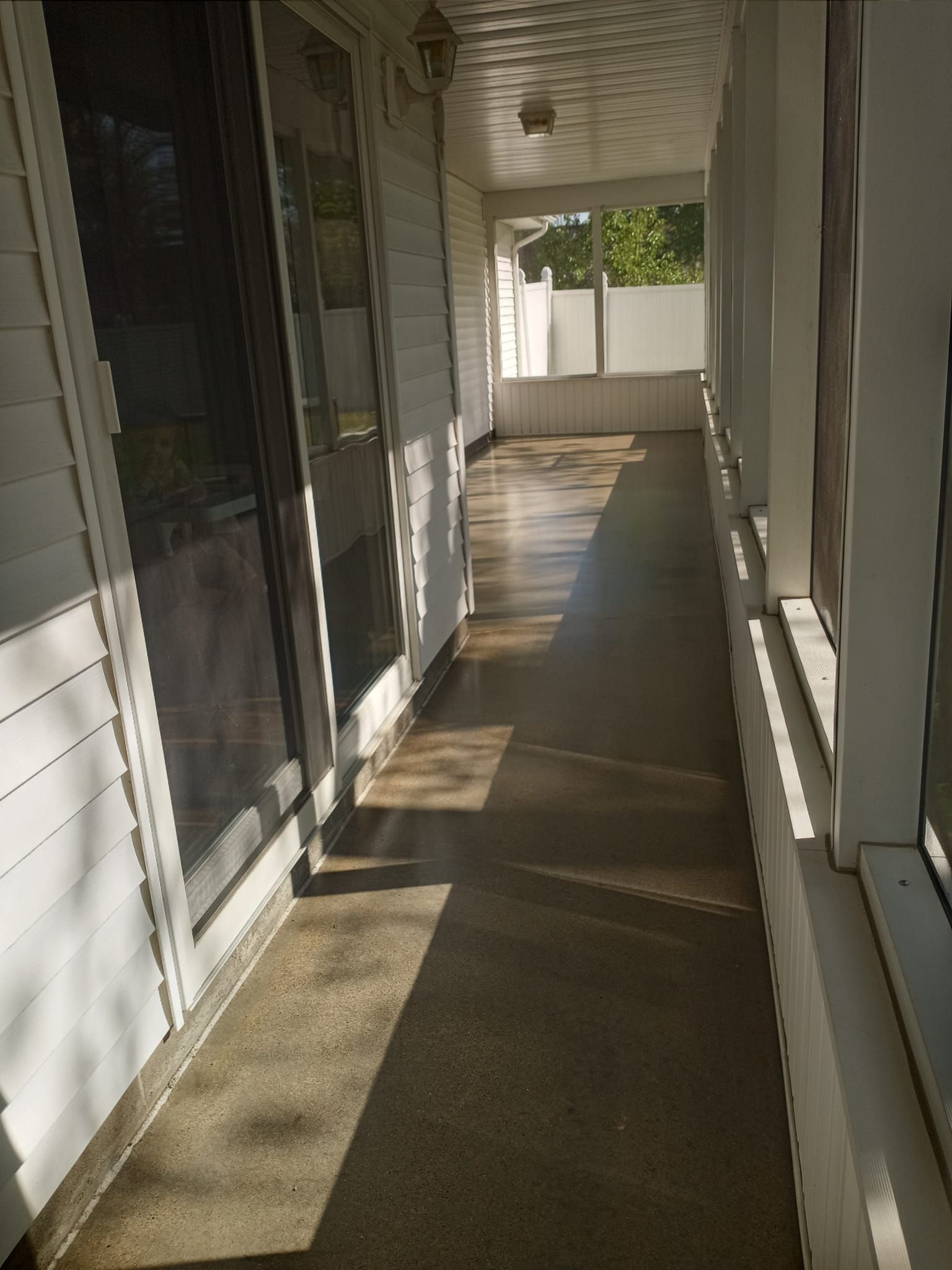 A sunlit, narrow covered porch with light siding, sliding glass doors, and screened windows along the side.