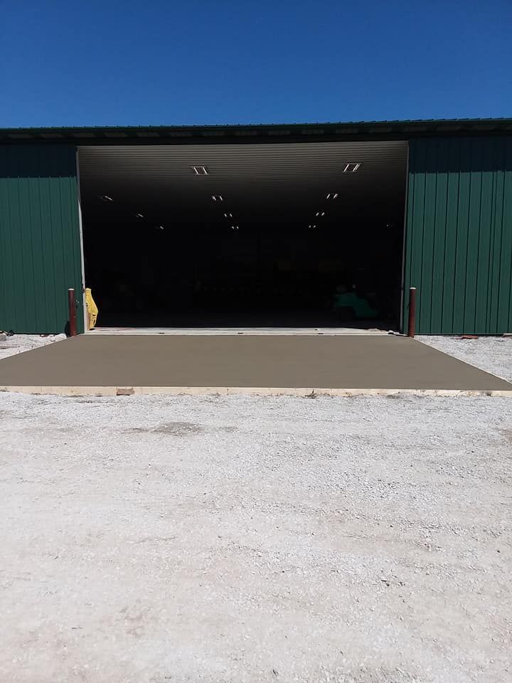 A green pole barn with a large open bay entrance and a freshly poured concrete apron sits above a gravel driveway.