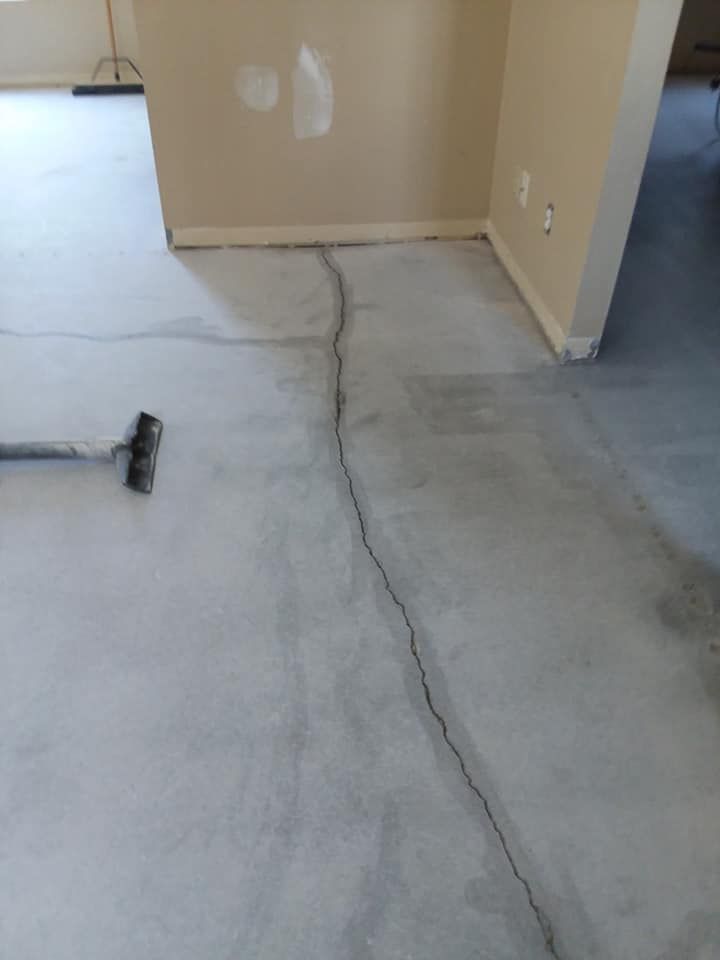 A long, thin crack runs through a bare concrete floor in a room with beige walls and a corner wall patch.