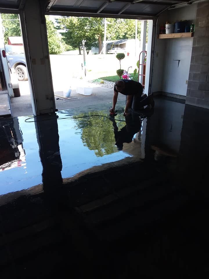 A person kneels on a garage floor while applying a dark, high-gloss coating that creates a clear reflection.