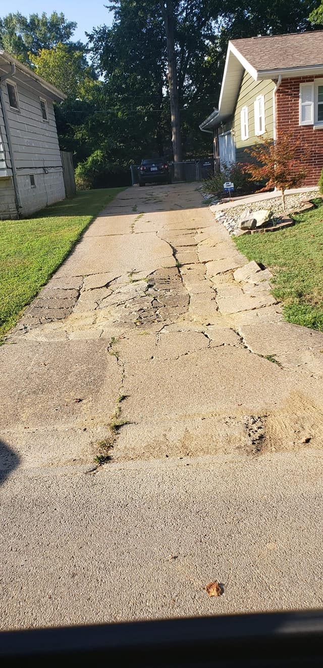 A severely cracked and weathered concrete driveway between two residential houses on a sunny day.