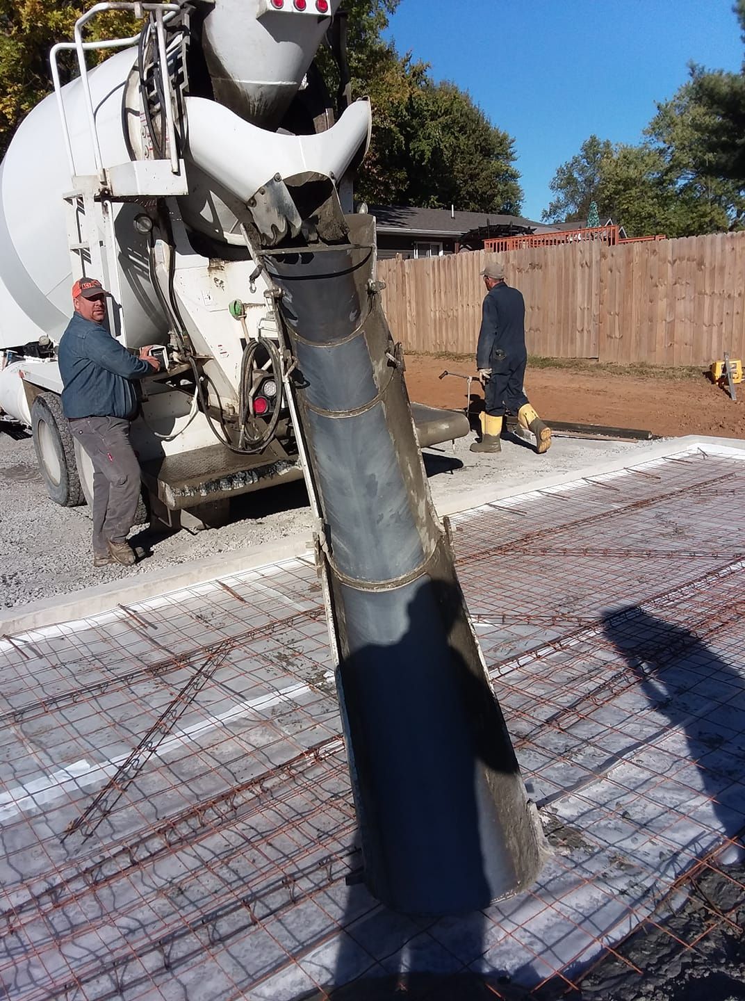 A concrete truck with a metal chute pours wet concrete onto a wire-mesh prepared site, with workers standing nearby.