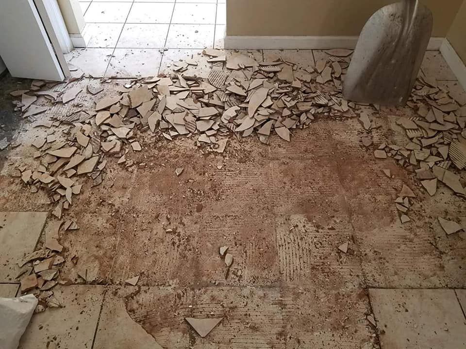 A pile of shattered beige ceramic floor tiles sits on a partially removed floor, with a metal shovel nearby.