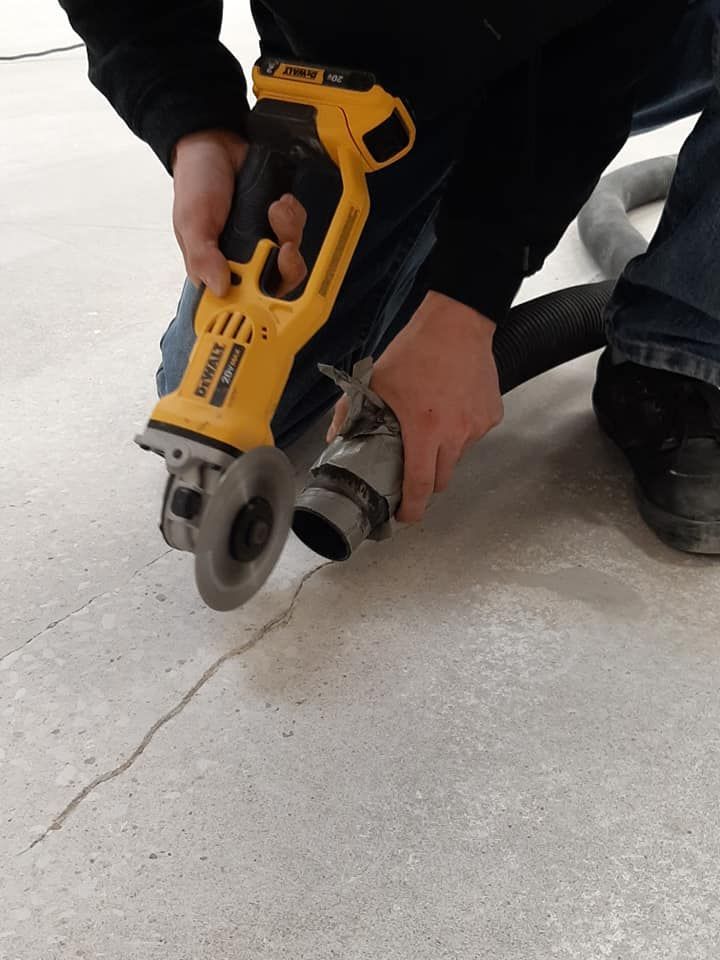 A person uses a yellow DeWalt angle grinder to cut a crack in a concrete floor while holding a vacuum hose for dust control.