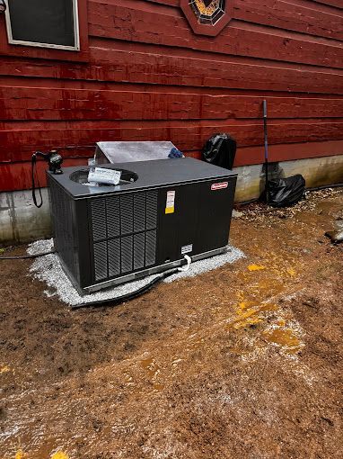 A black air conditioner is sitting on the ground in front of a red building.