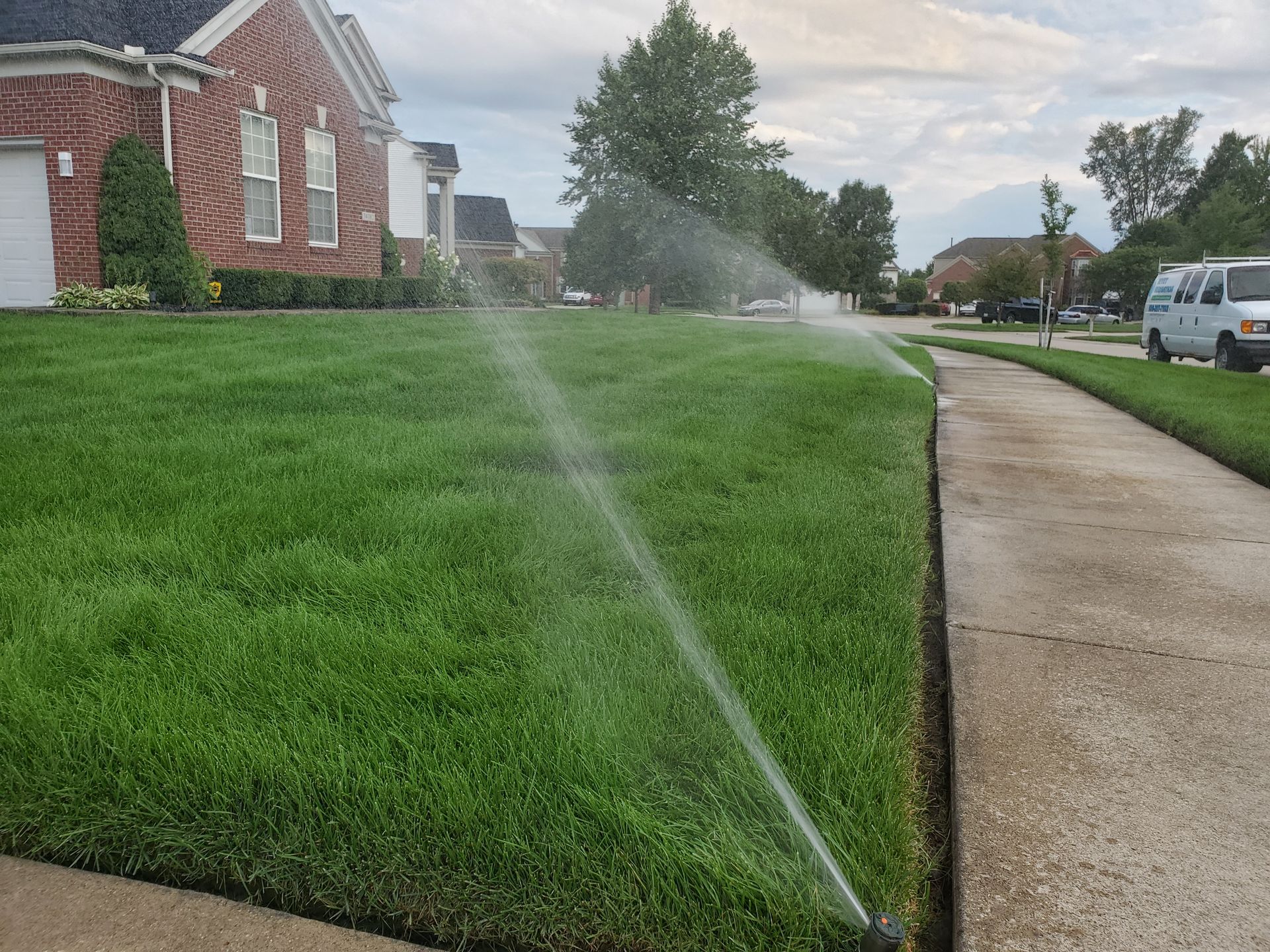 A lawn sprinkler is spraying water on a lush green lawn in front of a brick house.