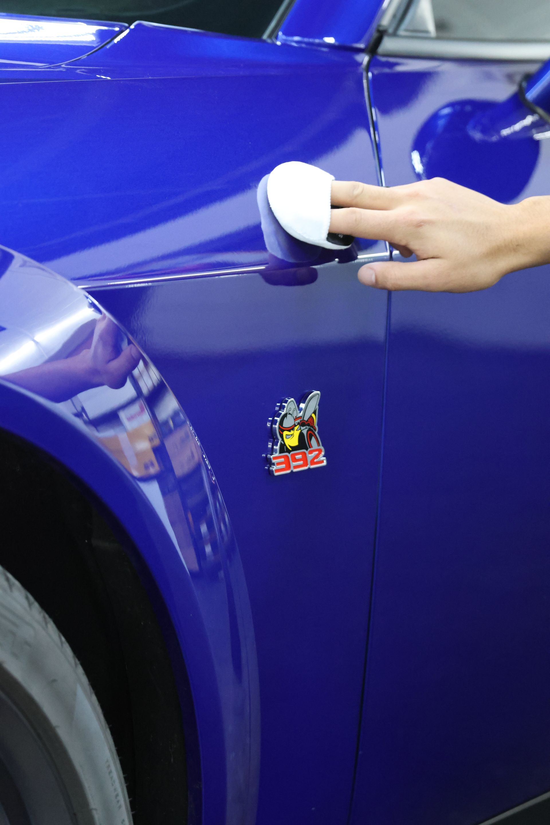 A person uses a white microfiber pad to apply product to the blue fender of a Dodge vehicle with a 392 Scat Pack emblem.