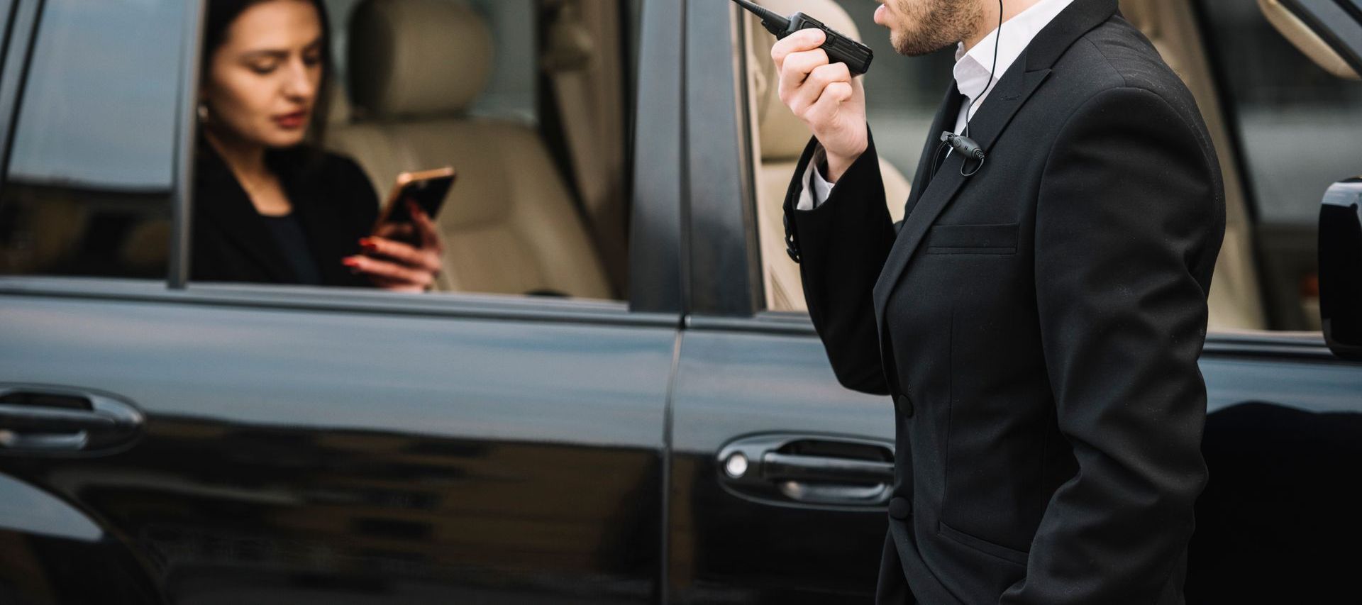 A security professional in a suit speaks into a two-way radio beside a car where a passenger checks a smartphone.