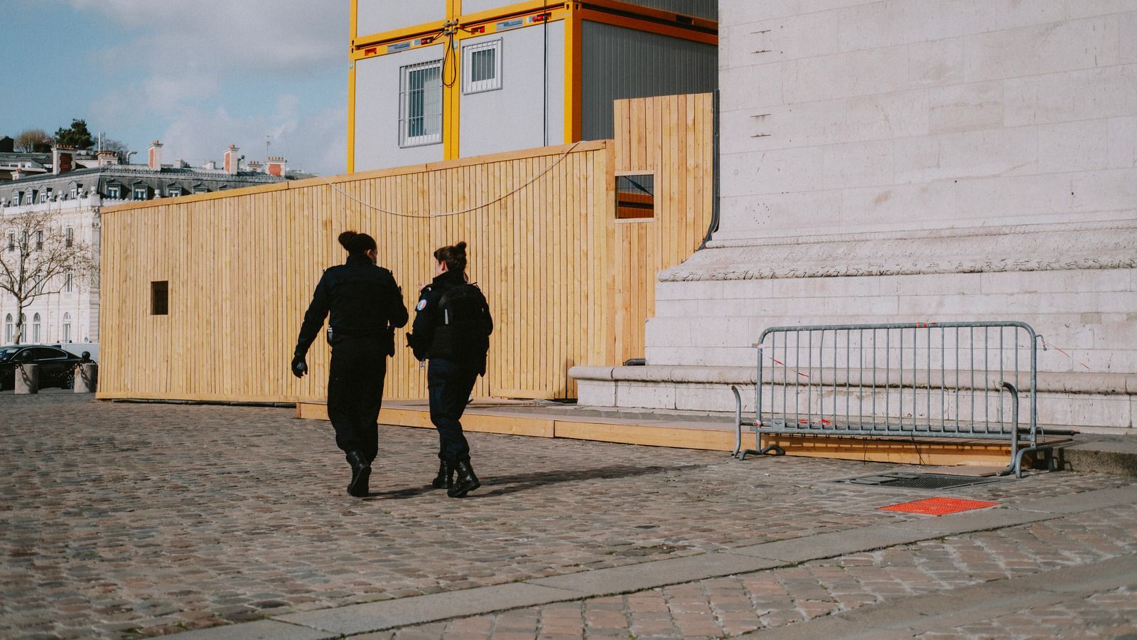 Two uniformed officers walk past a wooden barrier in a cobblestone square. Yellow containers are visible.