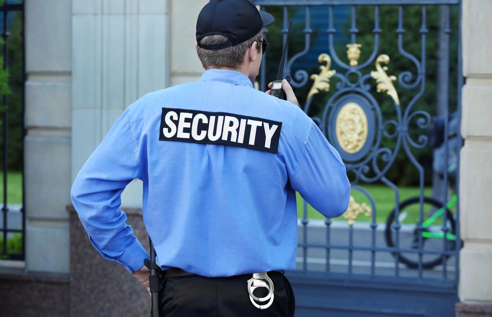 Security guard in blue shirt with “SECURITY” patch, standing near ornate gate, talking on radio.