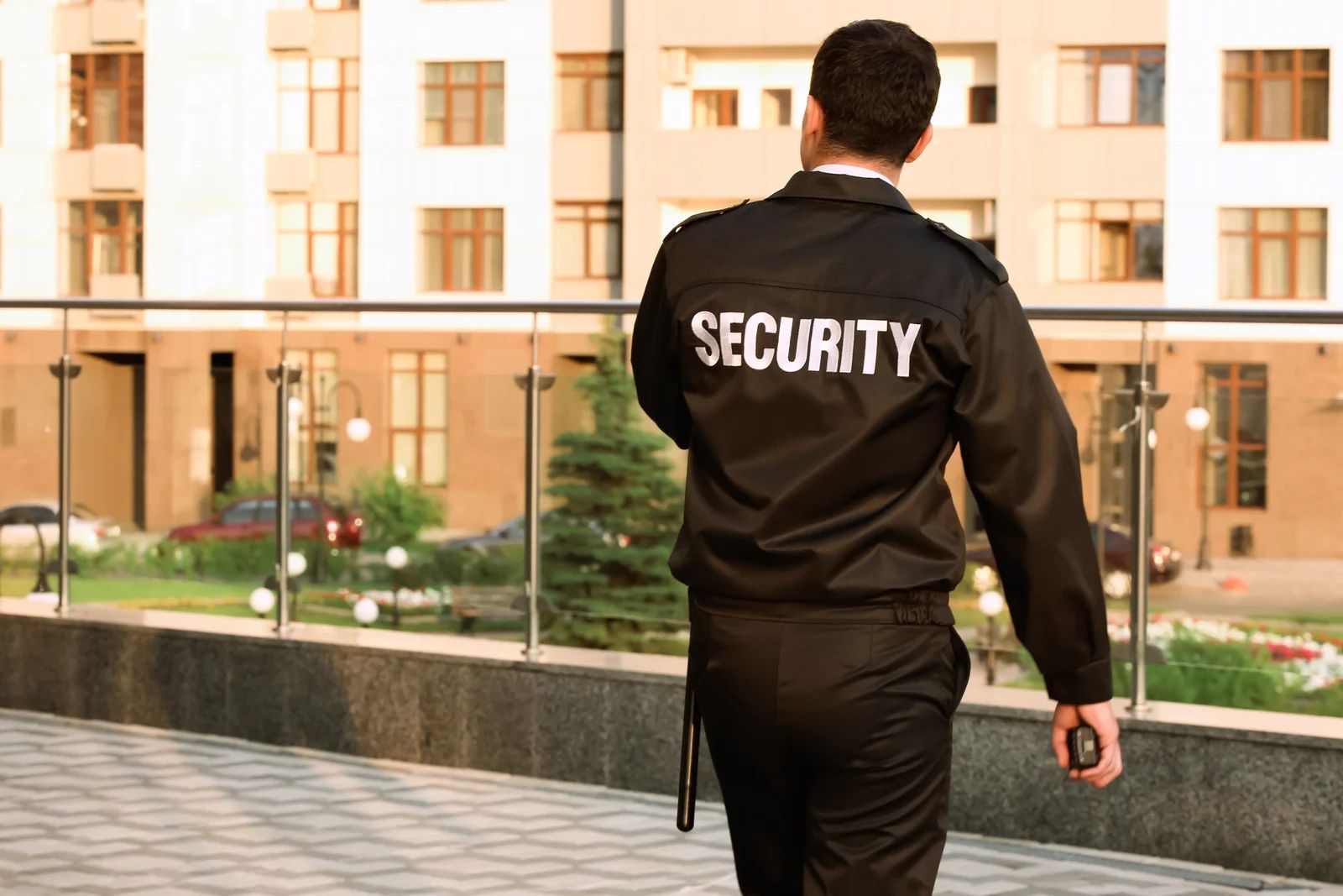 Security guard in black uniform walking outdoors, holding a radio. Building in background.