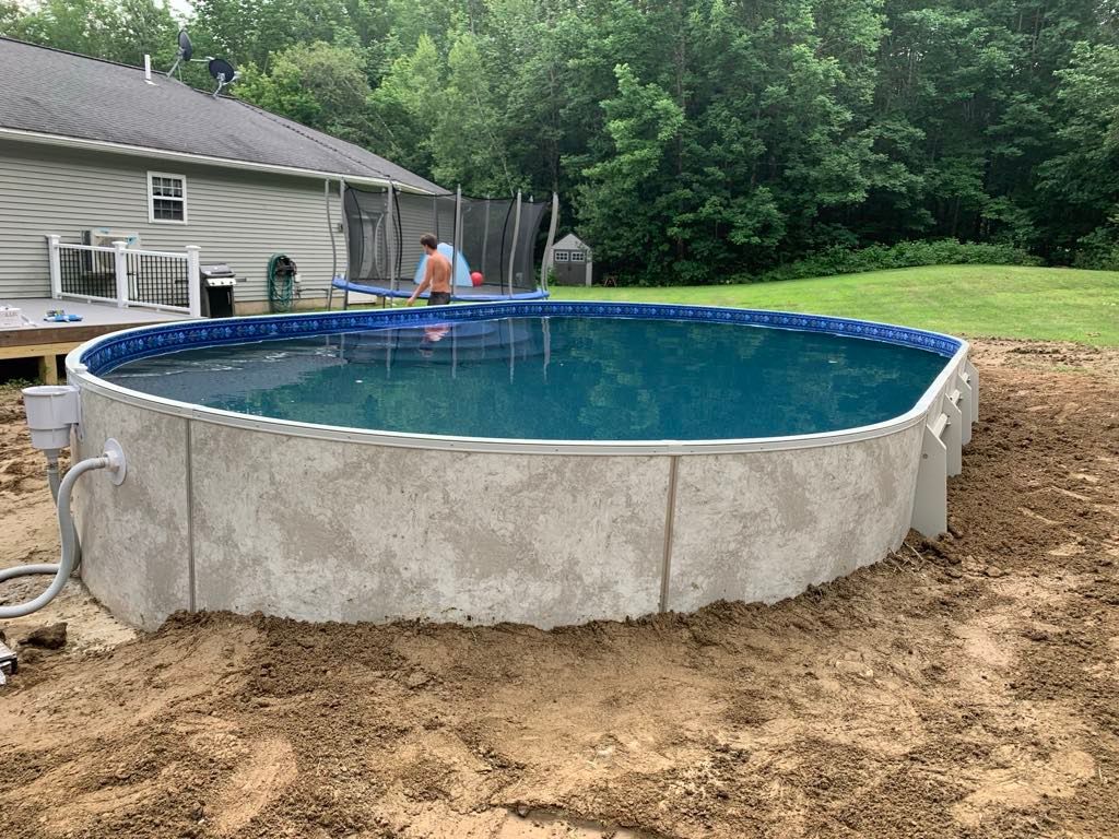 A large swimming pool is sitting in the dirt in front of a house.