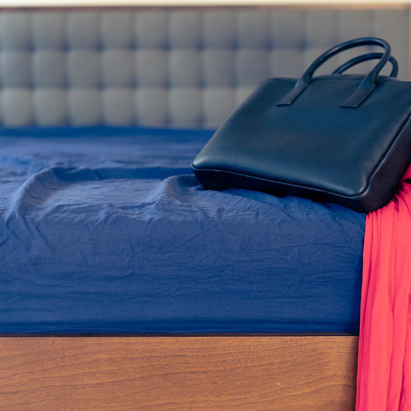 Blue handbag on a rumpled blue bedsheet with a red garment; wooden bed frame and upholstered headboard.