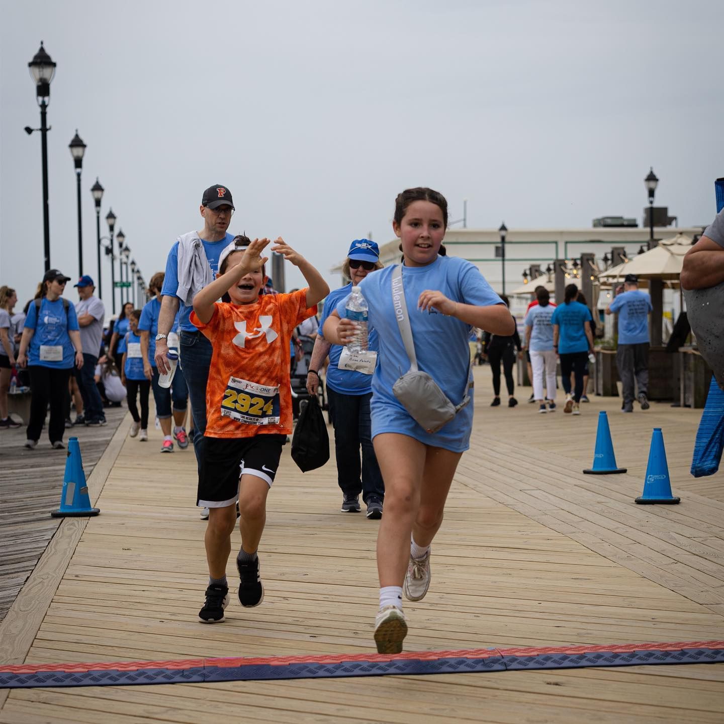 Two children running towards a finish line on a boardwalk, surrounded by people.
