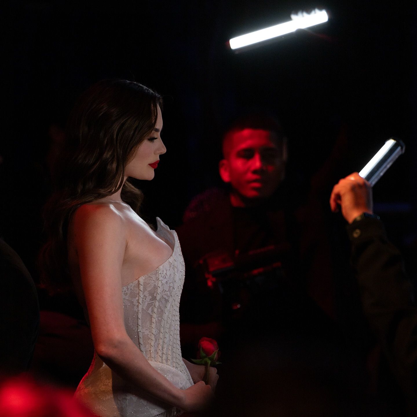 Woman in a white gown poses under stage lights, with a man holding a light in the background.