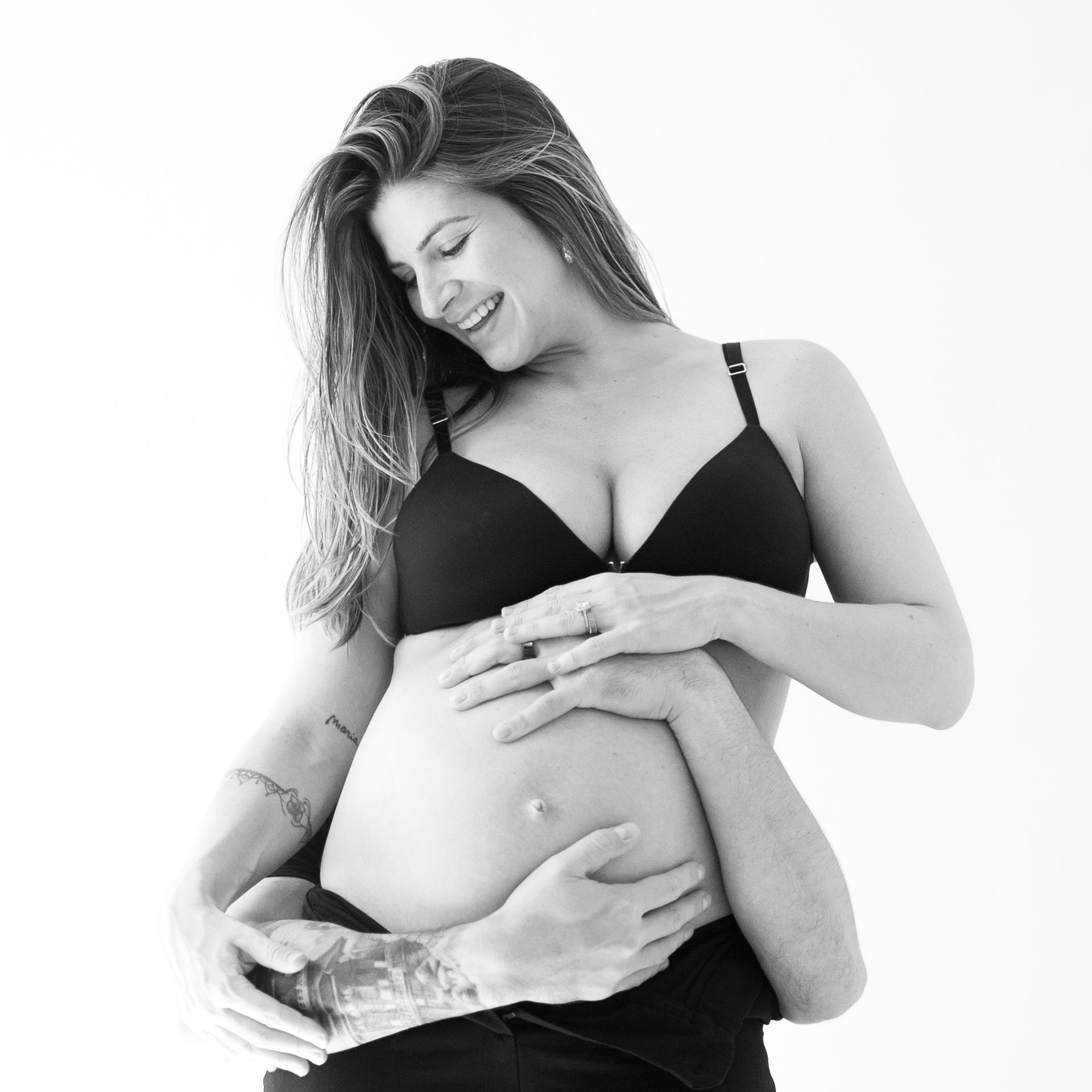 Pregnant person smiling, cradling belly with partner's arms, black bra.