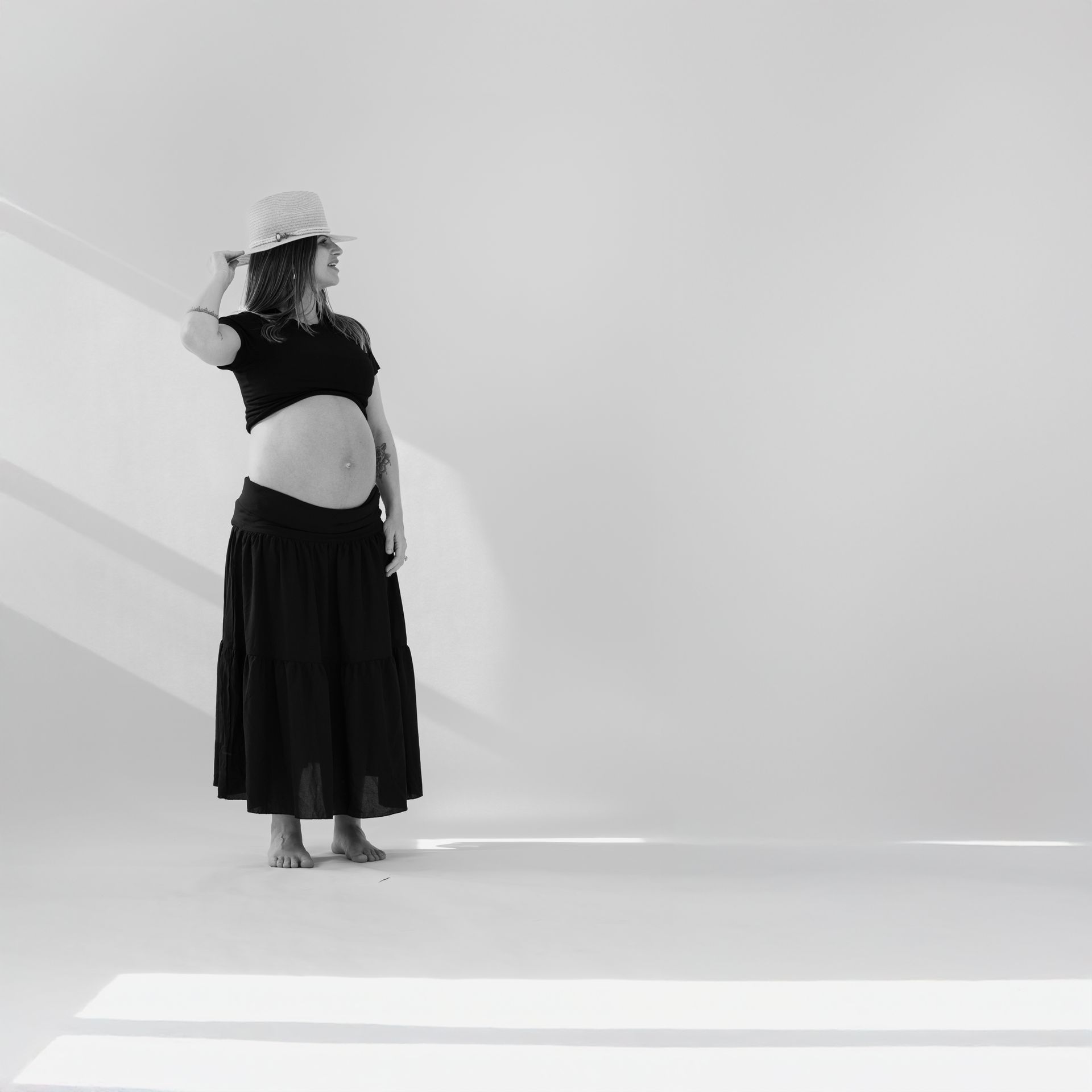Pregnant person in black outfit and hat, hands on hat, looking away in a studio setting.