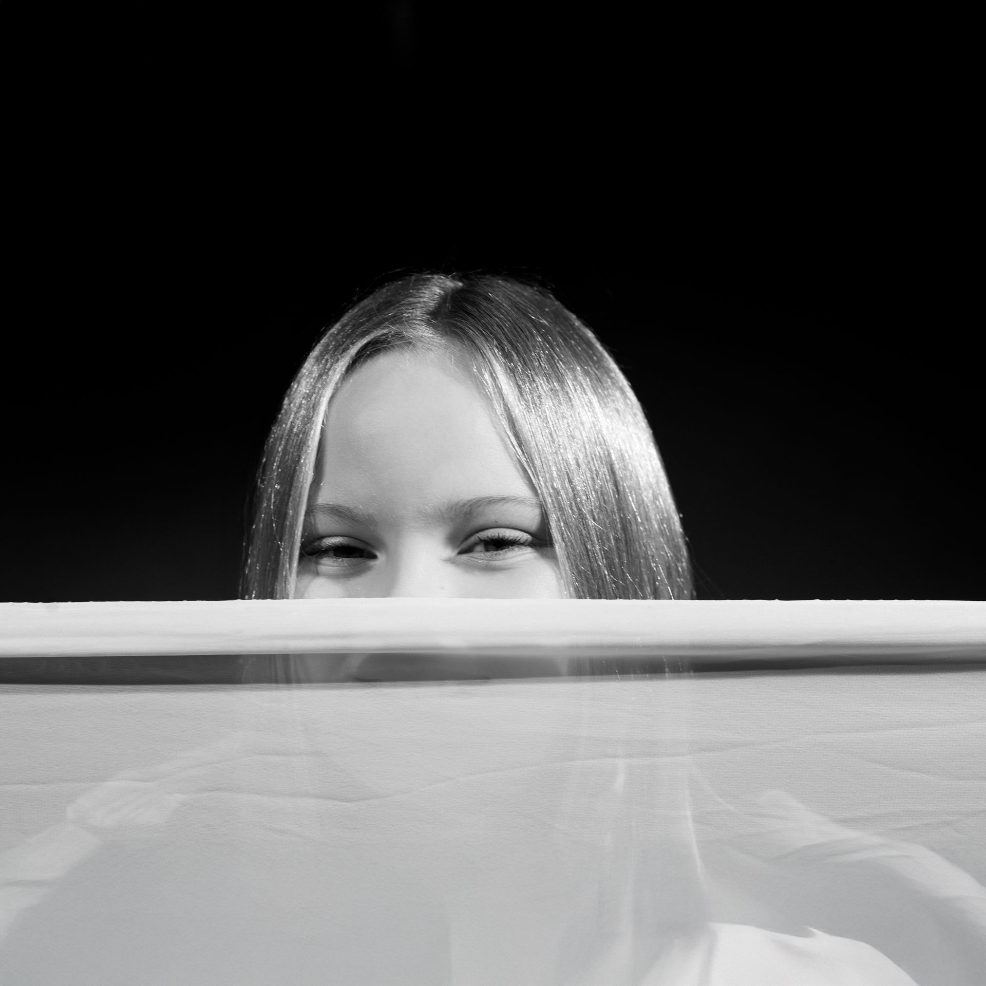 Blond-haired person peering over a white surface with a slight smile, against a dark background.