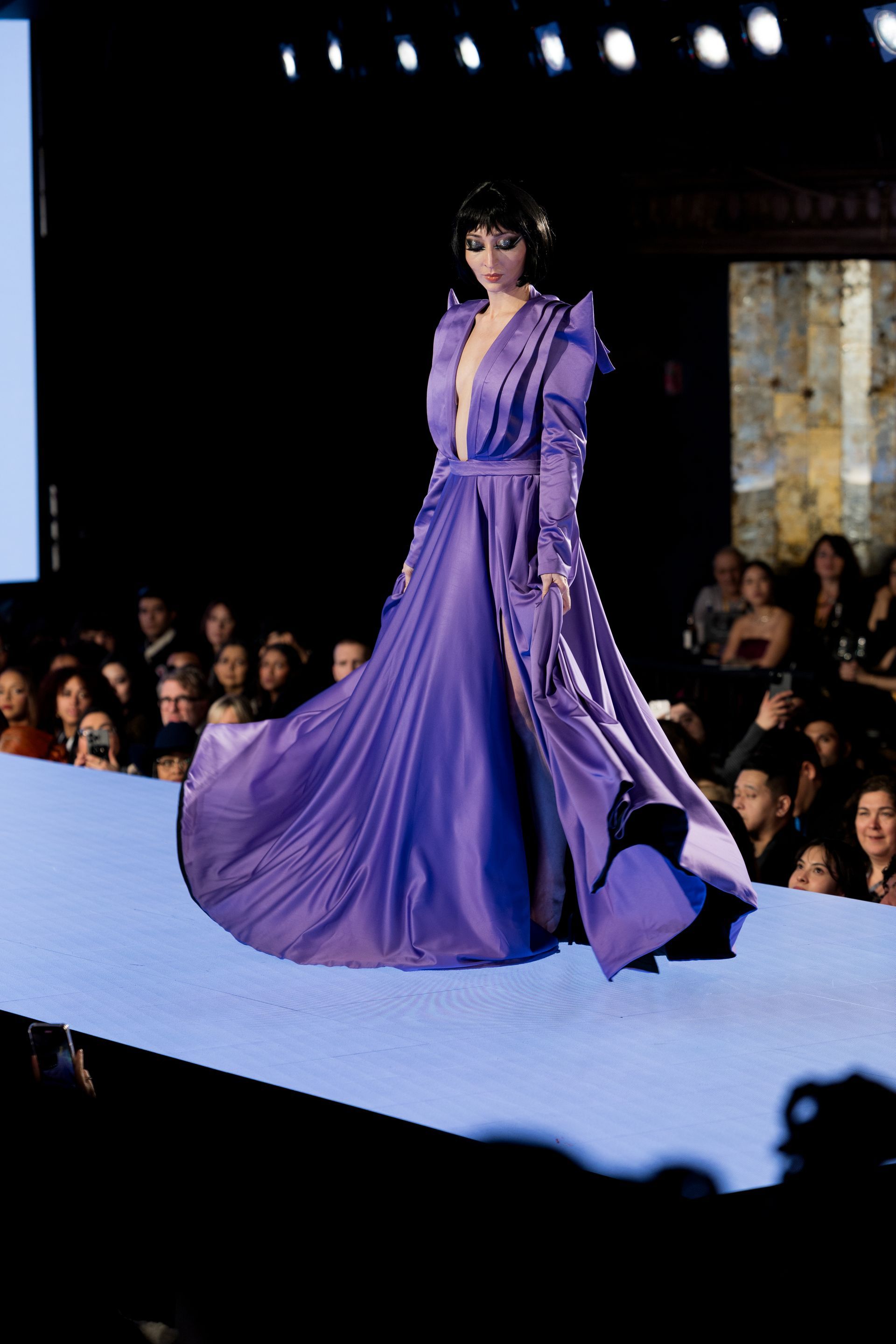 Model in a long, flowing purple gown walks a runway. Deep neckline, shoulder detail, and audience in background.