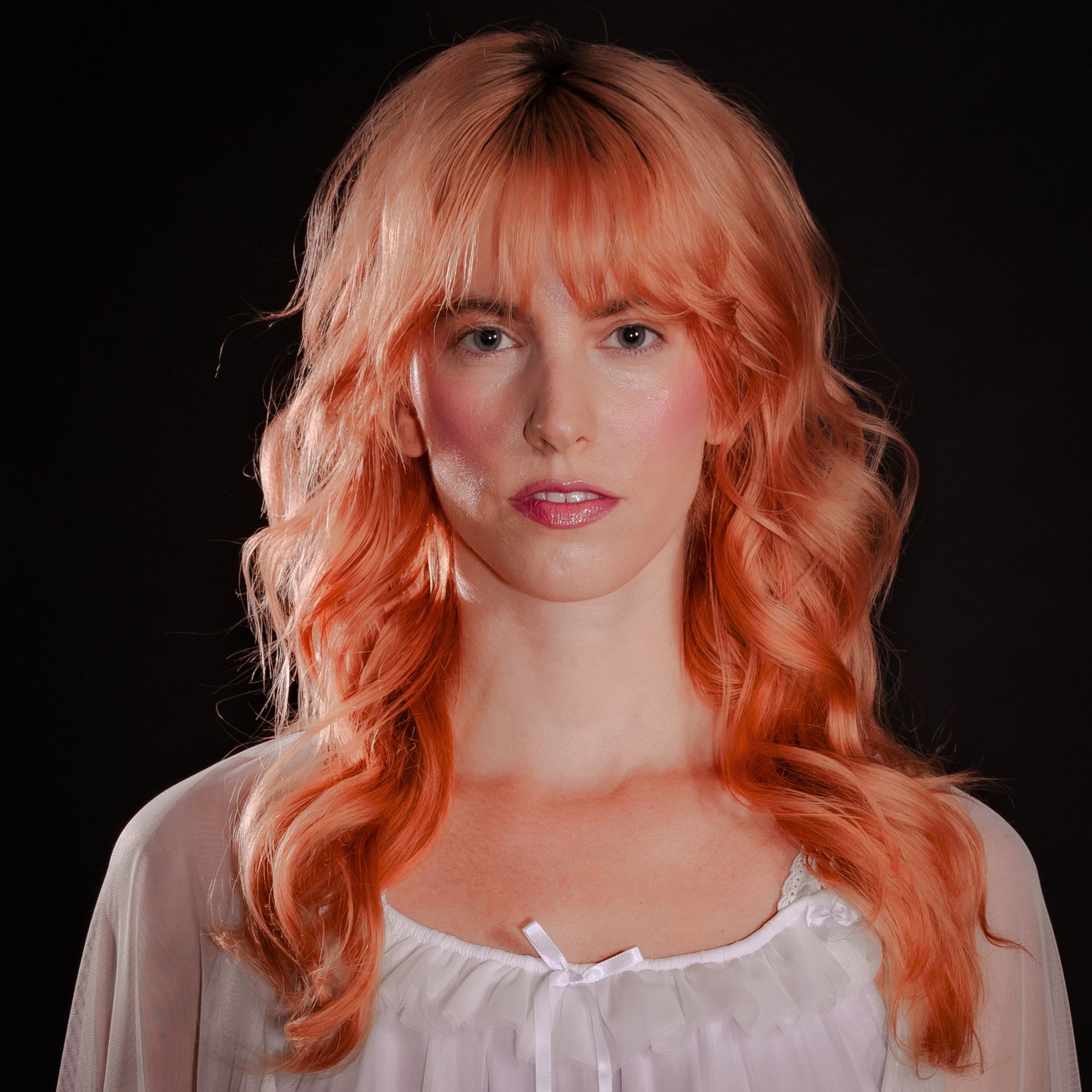Woman with pinkish-orange hair and a neutral expression against a black background. She wears a white top.