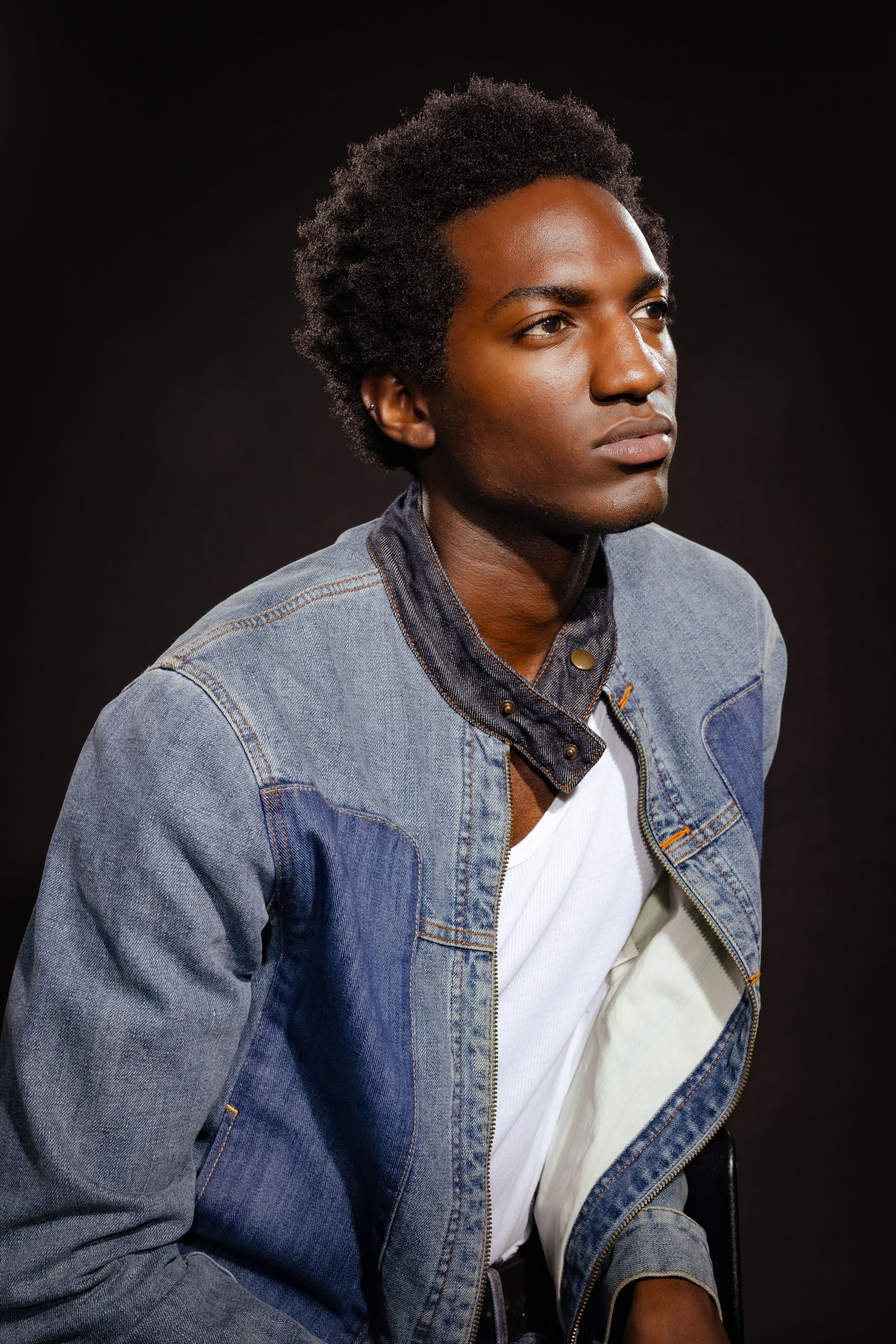 Man wearing denim jacket and white shirt, looking off-camera against a black backdrop.