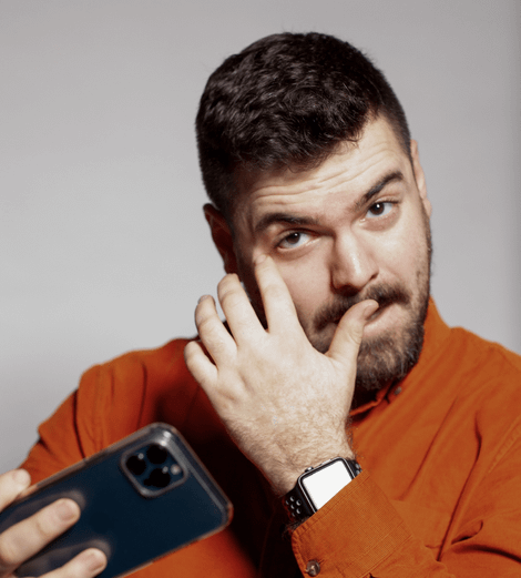 Man in orange shirt with phone, touching face, looking at the camera with a doubtful expression.