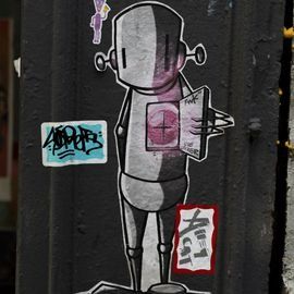 Robot sticker with pink center and open chest, on a dark post with graffiti stickers.