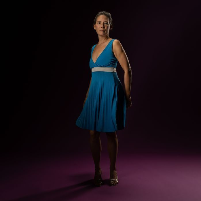 Woman in teal dress, hands behind back, standing on purple floor against a dark background.