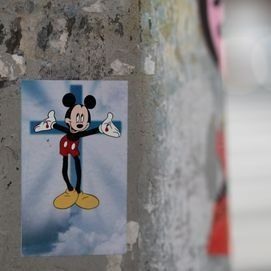 Mickey Mouse on a cross sticker on a textured surface, arms outstretched with a cloudy background.