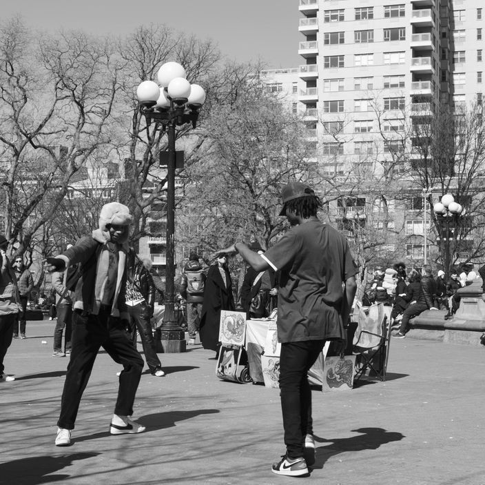 Two people dancing in a park, near a street vendor stall and buildings.