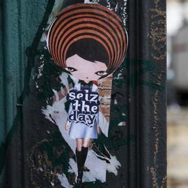 A street art sticker of a cartoon girl with striped head, holding a sign that says