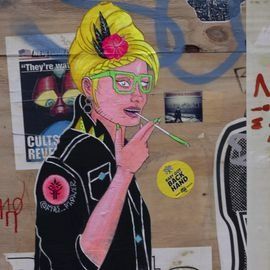 Street art of a person with blonde hair and green glasses holding a cigarette, making a peace sign.